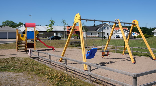 Fotografiet är en översiktsbild över Äppelkullens lekplats. I förgrunden syns en gul gungställning med tre platser. I bakgrunden, till vänster, syns en lekställning med en rutschkana. Marken är sand och runt gungställningen är det staket. 