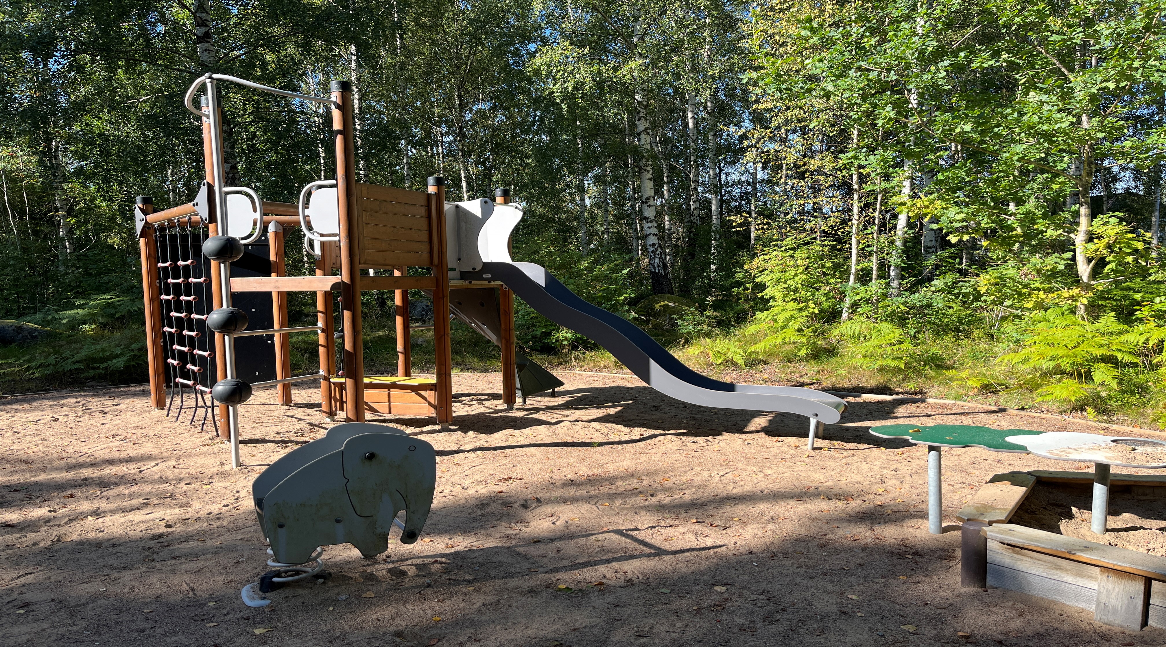 Foto av en lekplats som ligger i en solig skogsglänta. Underlaget är sand och på lekplatsen finns en klätterställning med rutschkana och ett gungdjur i form av en elefant.