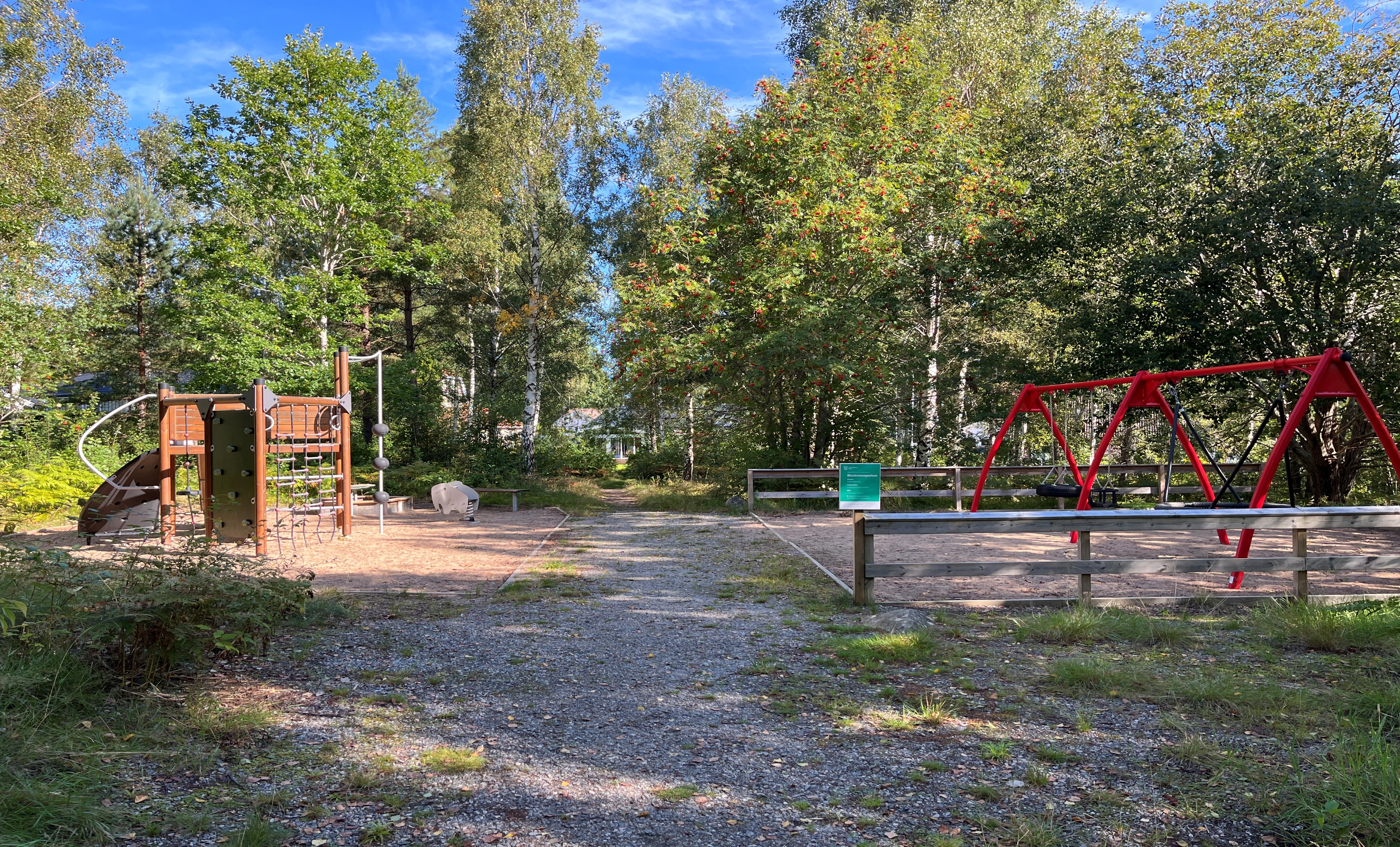 Foto av en tvådelad lekplats med en grusgång emellan. Lekplatsen ligger i en skogsdunge. Till höger finns en röd gungställning för fyra gungor och till vänster en klätterställning och ett gungdjur i form av en elefant.