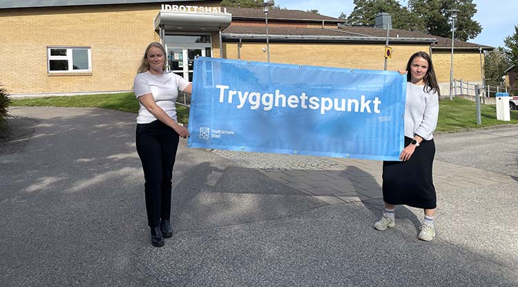 Beredskapsstrateg Victoria Johnson och säkerhetssamordnare Olivia Wernberg framför idrottshallen på MÅG med blå vepa som har texten “Trygghetspunkt” 