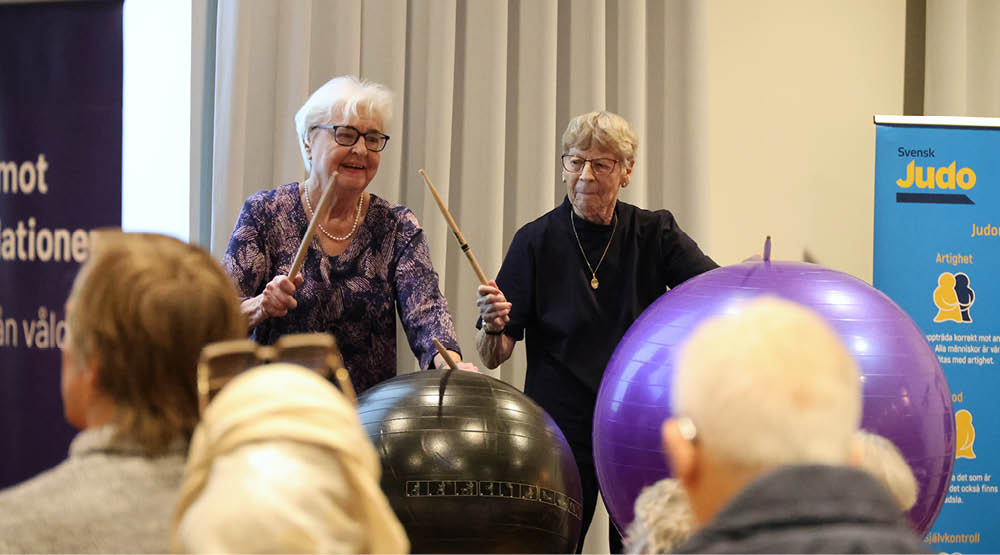 Två damer som håller i trumpinnar och slår på en pilatesboll.