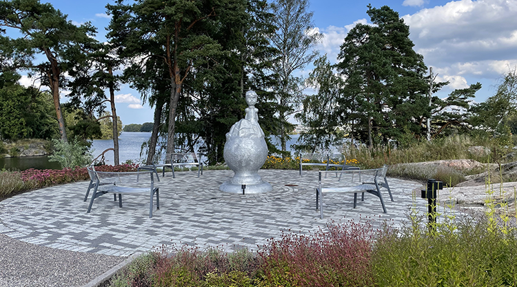 Skulptur av blank aluminium omgiven av bänkar. Skulpturen föreställer ett barn med pilothjälm, den har puffärmar och kikar ut från ett berg på toppen av ett klot. 